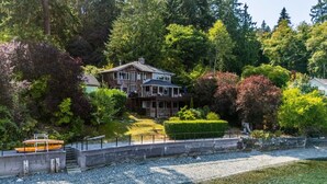 Une maison à deux étages avec un quai et un bateau, entourée d’une forêt dense et offrant une vue dégagée sur l’eau.