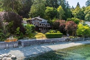 Ein zweistöckiges Haus mit einem Steg und einem Boot, umgeben von dichtem Baumbestand und freiem Blick auf das Wasser.