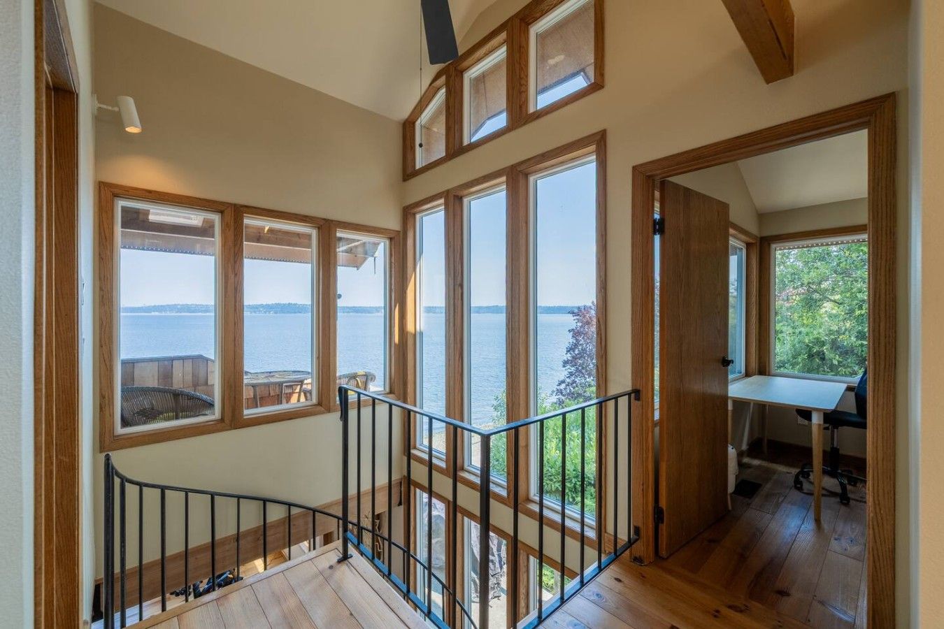 A wooden interior with large windows overlooking a body of water, a staircase with a metal railing, and a small table with a chair.