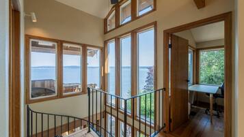 A wooden interior with large windows overlooking a body of water, a staircase with a metal railing, and a small table with a chair.