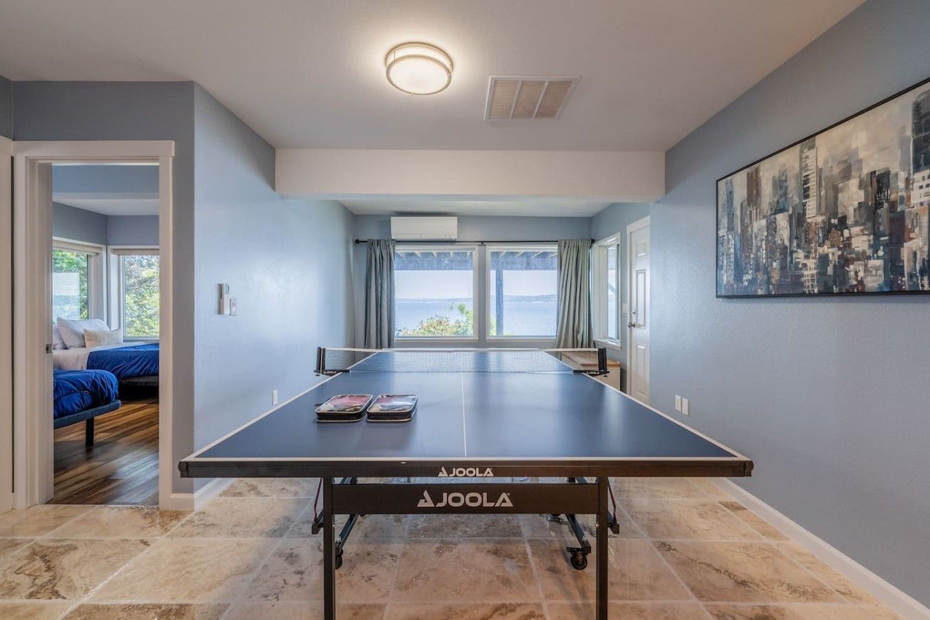 A room with a table tennis table, a bed with blue bedding, and a cityscape painting on the wall.