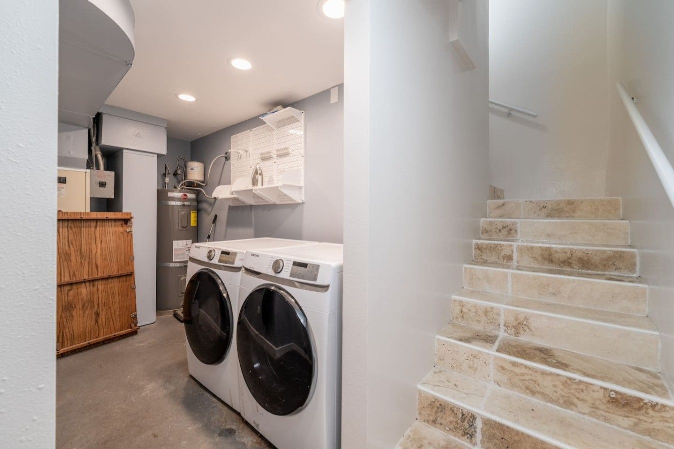 A laundry area with two front-loading washing machines, a wooden cabinet, and a staircase with beige tiles.