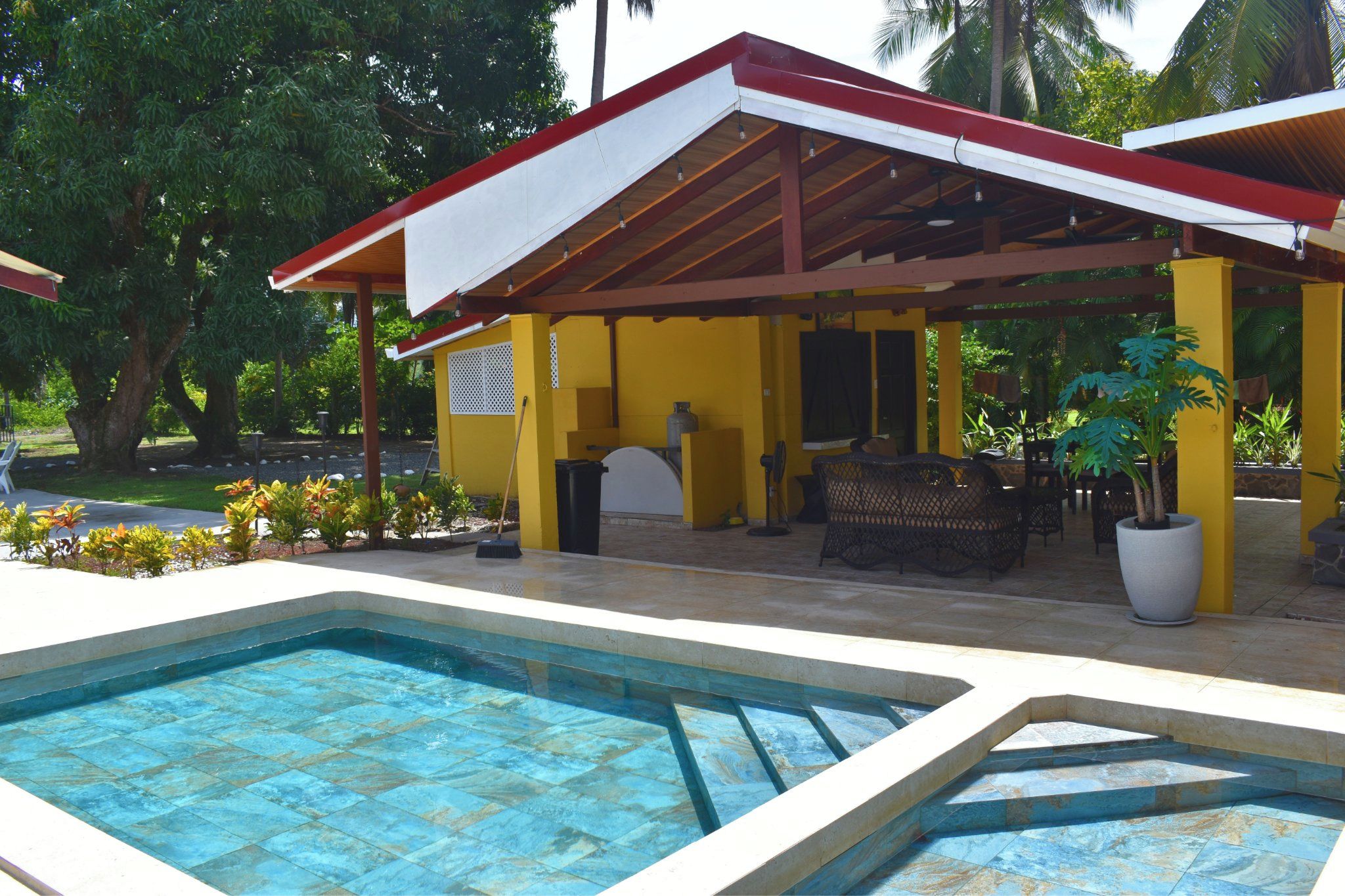 Un área de piscina con una zona de estar cubierta y un edificio amarillo al fondo.