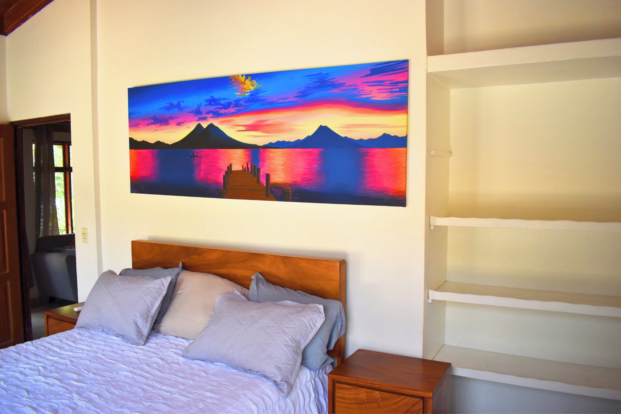 Un dormitorio con cama, almohadas, cabecero de madera, mesita de noche y un cuadro colgado en la pared con un atardecer sobre el agua.