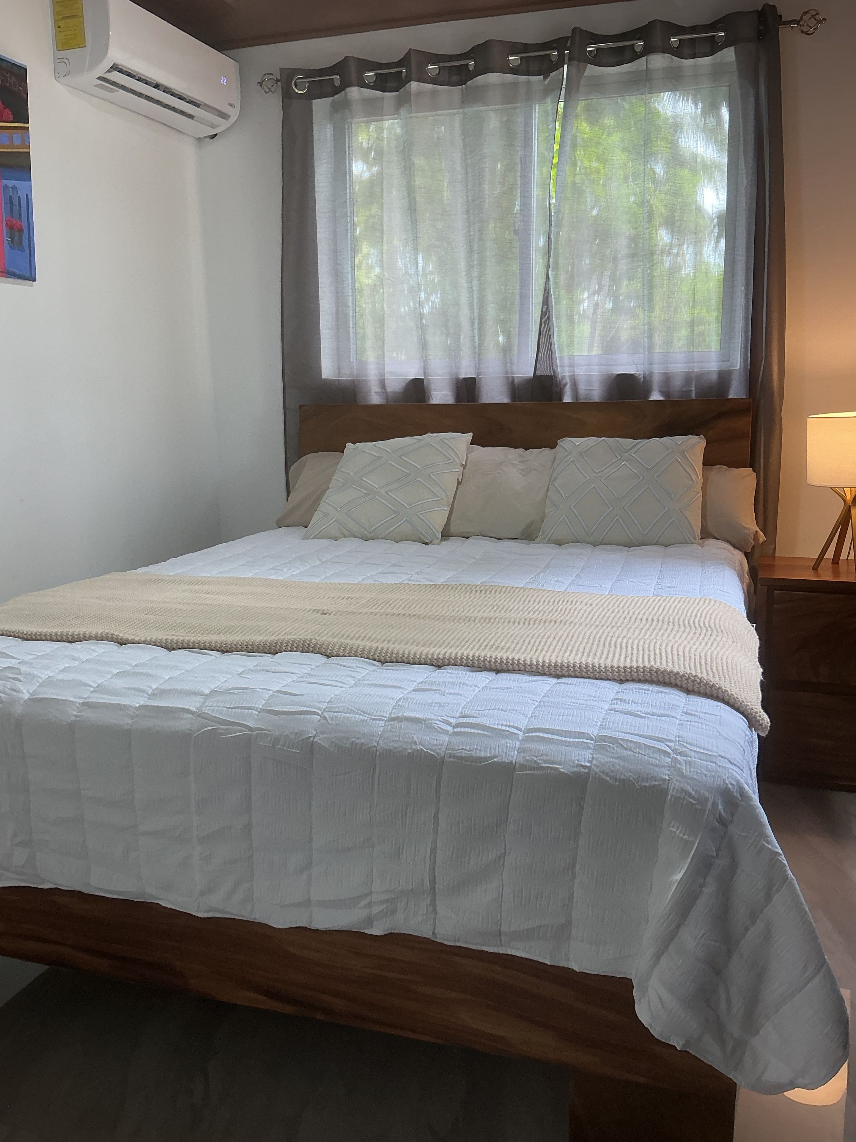 Una cama con colcha blanca acolchada, cabecera de madera y una ventana con cortinas traslúcidas.