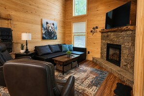 A cozy cabin interior with a stone fireplace, a sofa, a coffee table, and a television. - NEW CABIN! - MawMaw Bear's Nook -King Bed-Dog Yard (Murphy)