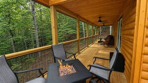 En overdækket terrasse med bålpit-bord og stole, omgivet af skov.
