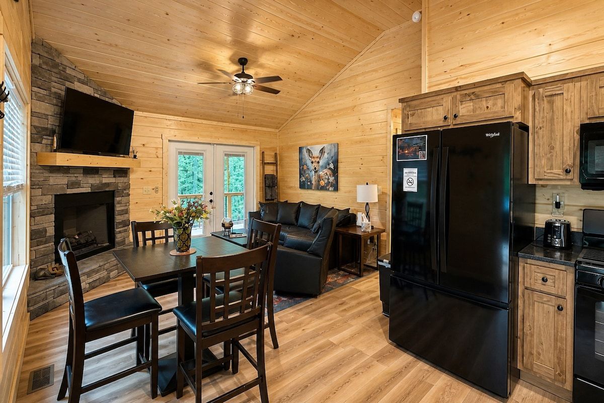 A wooden cabin interior with a dining area, a fireplace, a black refrigerator, and a ceiling fan.
