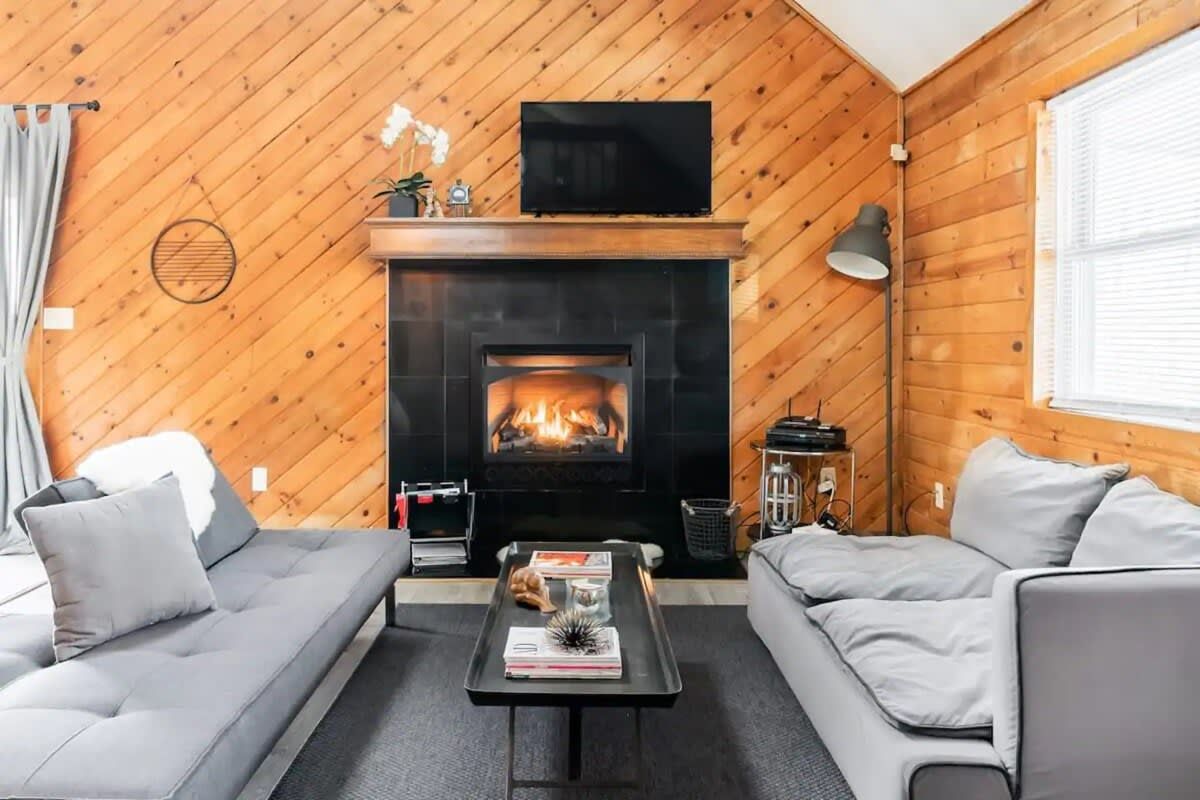 A cozy living room with a fireplace, two grey sofas, a coffee table, and a TV on a wooden wall.