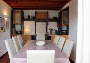 A dining area with a table set for six, surrounded by chairs, in a room with wooden beams on the ceiling and a kitchen area in the background. - Waterville Holiday Home (Kerry)