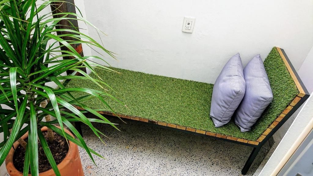 Un banco con asiento de césped artificial verde y dos cojines, una planta en maceta a la izquierda y una pared blanca con toma de corriente.