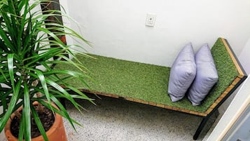 Un banco con asiento de césped artificial verde y dos cojines, una planta en maceta a la izquierda y una pared blanca con toma de corriente.