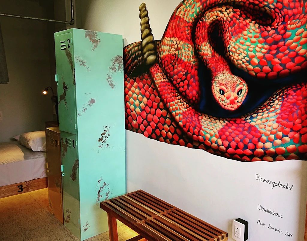 Una habitación con una cama, un armario metálico verde, un banco de madera y un mural grande de una serpiente en la pared.