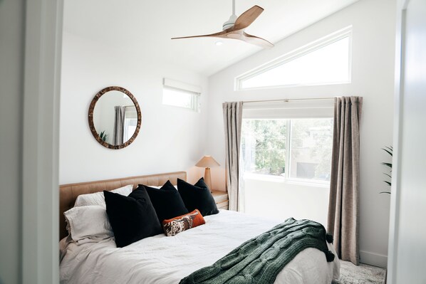 Ein Schlafzimmer mit einem Bett, einem Deckenventilator, einem Fenster mit Vorhängen und einem Spiegel an der Wand.