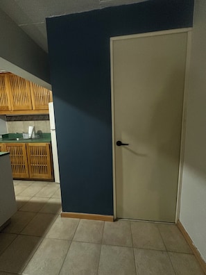 A room with a closed door, a kitchen area with wooden cabinets, and a tiled floor.