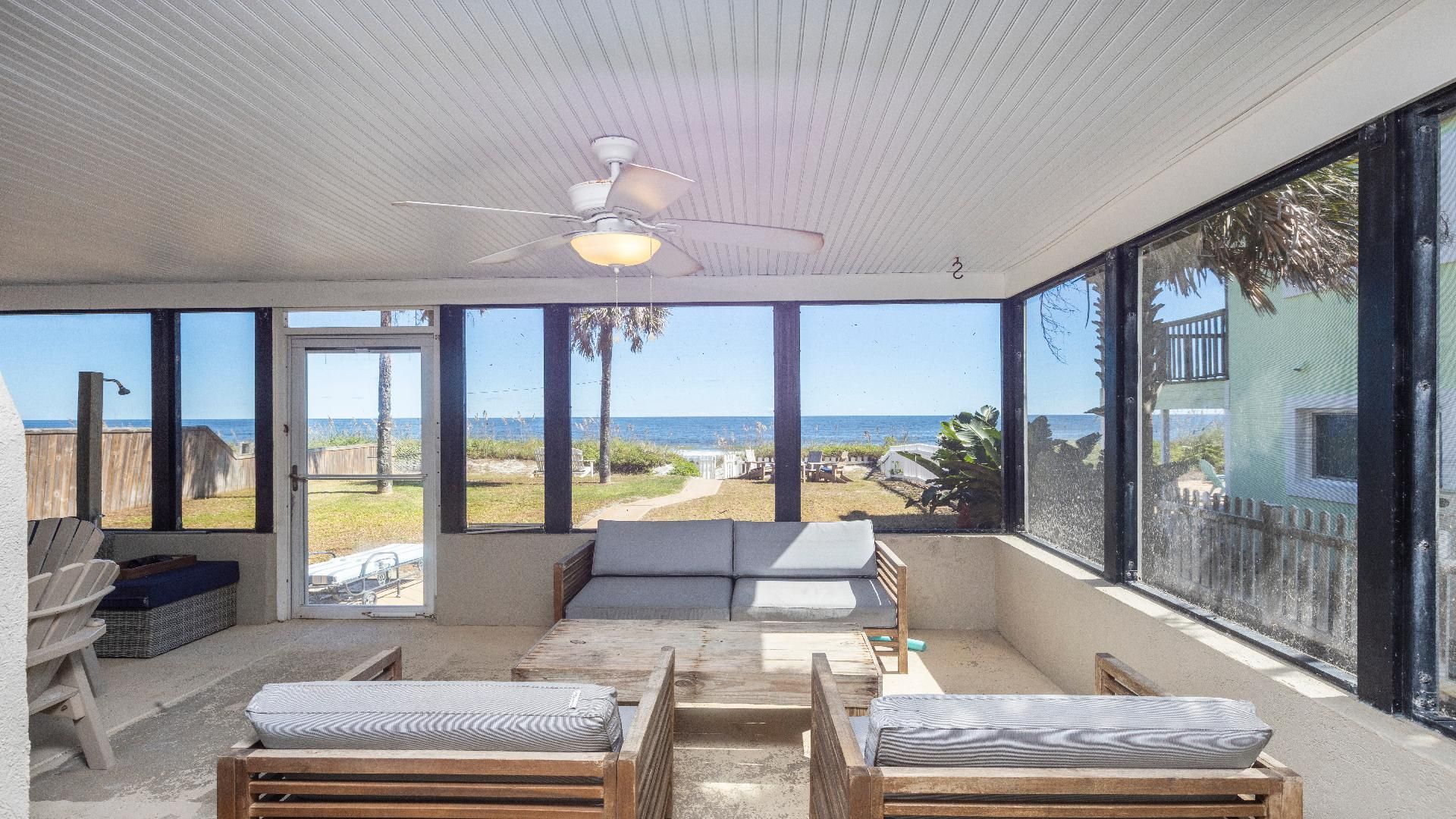 Una habitación iluminada por el sol con ventilador de techo, amplios ventanales con vista a la playa y una zona de estar con cojines.