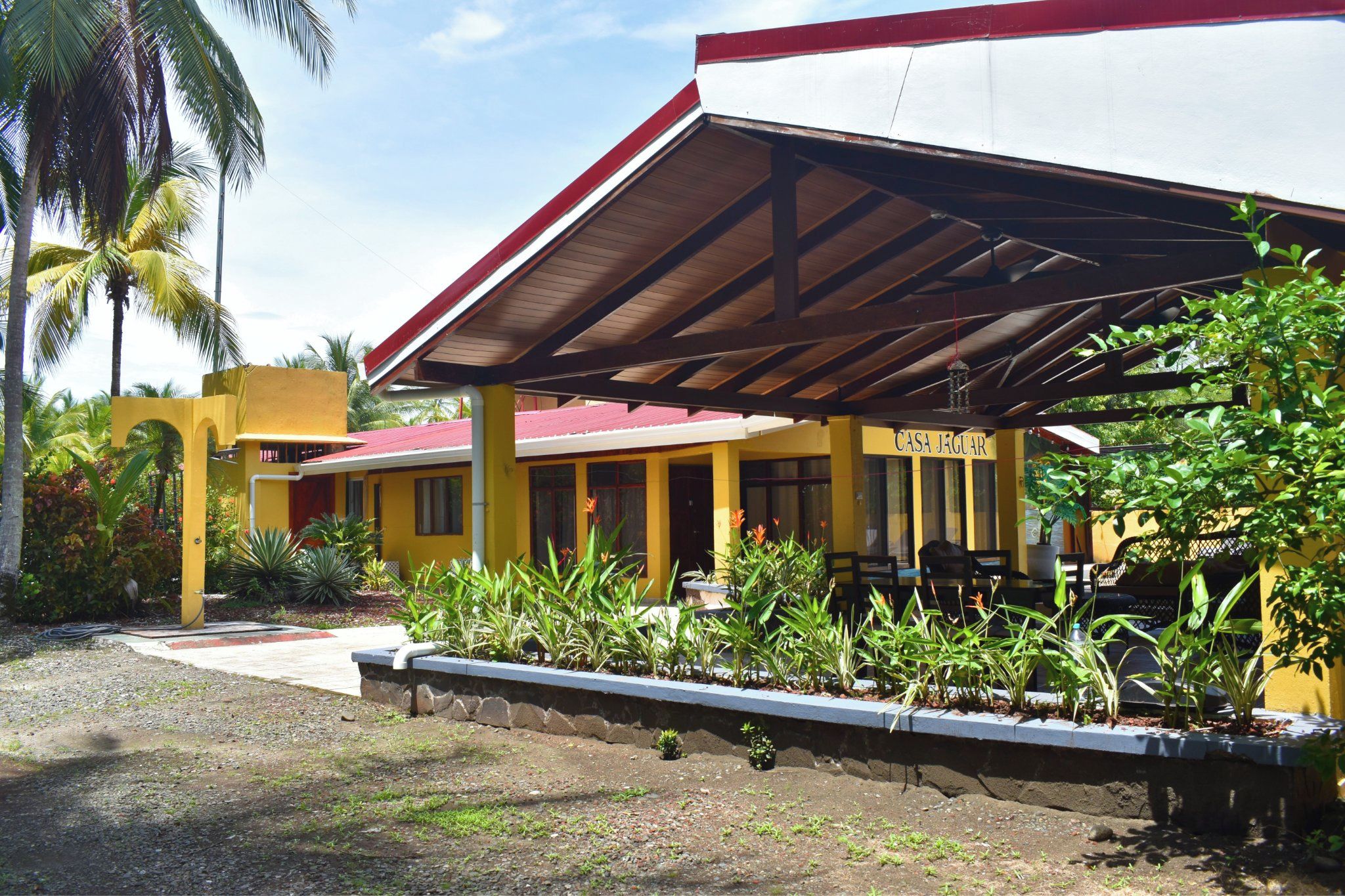 Un bâtiment jaune avec un toit rouge, portant l’inscription « CASA JAGUAR », entouré de plantes tropicales et de palmiers.
