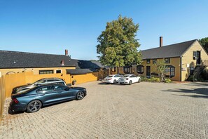 A parking area with two cars, a building with a sloped roof, and a large tree.