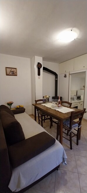 En kompakt opholdsstue med en sofa, et spisebord dækket med glas og en flaske, samt et tekøkken i baggrunden.