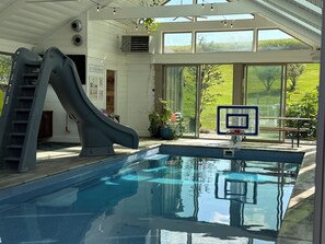 An indoor swimming pool with a slide, a basketball hoop, and large windows offering a view of the outdoors. - Bloom On The Spring: A modern Retreat in Nature. (Chambersburg)