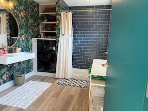 A bathroom with a white sink, a mirror with a floral frame, a black washing machine, a shower with a beige curtain, and a dark brick wall. - Bloom On The Spring: A modern Retreat in Nature. (Chambersburg)