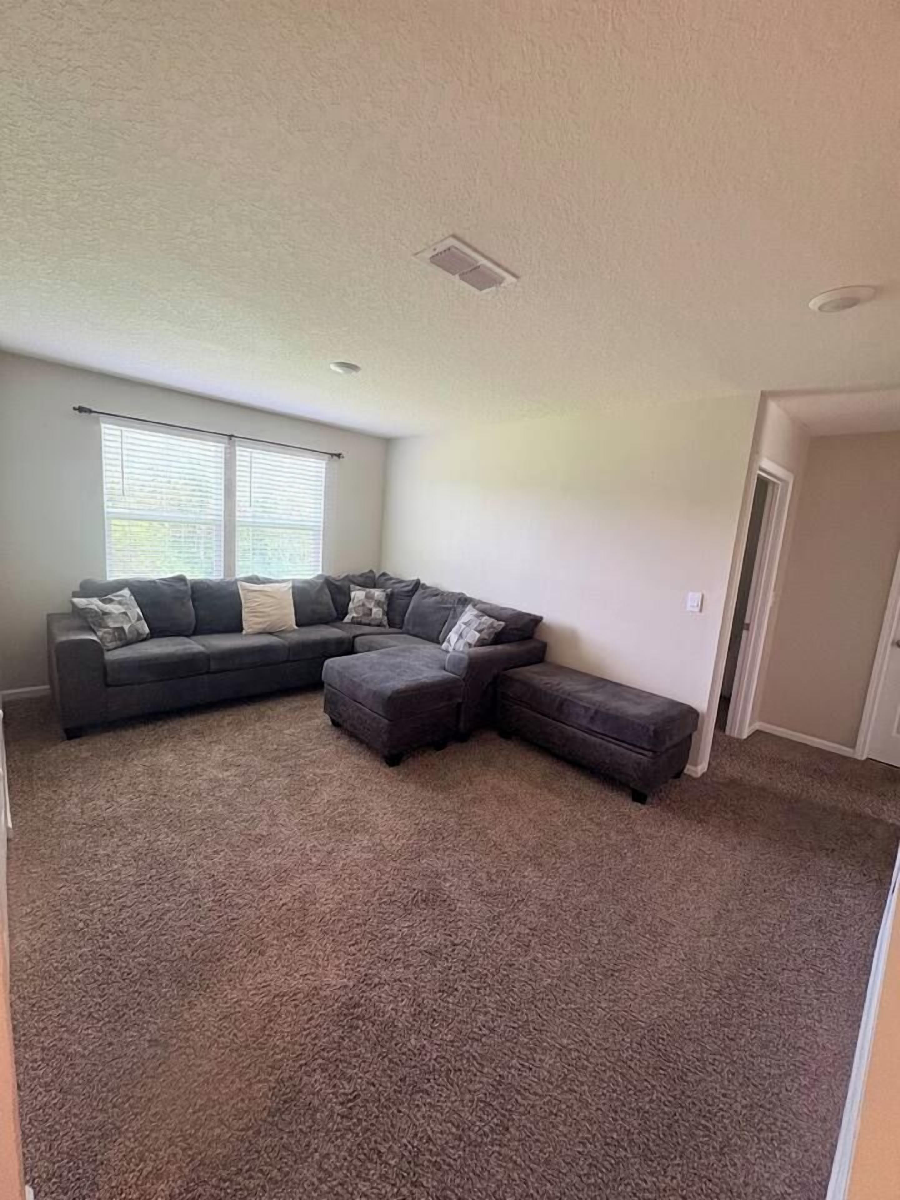 A living room with a sectional sofa, ottoman, and a window with blinds.