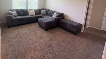 A living room with a sectional sofa, ottoman, and a window with blinds.