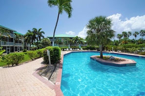 A swimming pool with a surrounding walkway, palm trees, and a building with a green roof. - Beach Living at Plantation Village BLD (George Town)