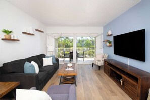 A modern living room with a black sofa, a coffee table, a TV, and a balcony with a view of palm trees. - Beach Living at Plantation Village BLD (George Town)