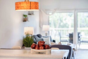 Un coin repas avec une table blanche, un saladier de pommes rouges, une plante en pot et une lampe.