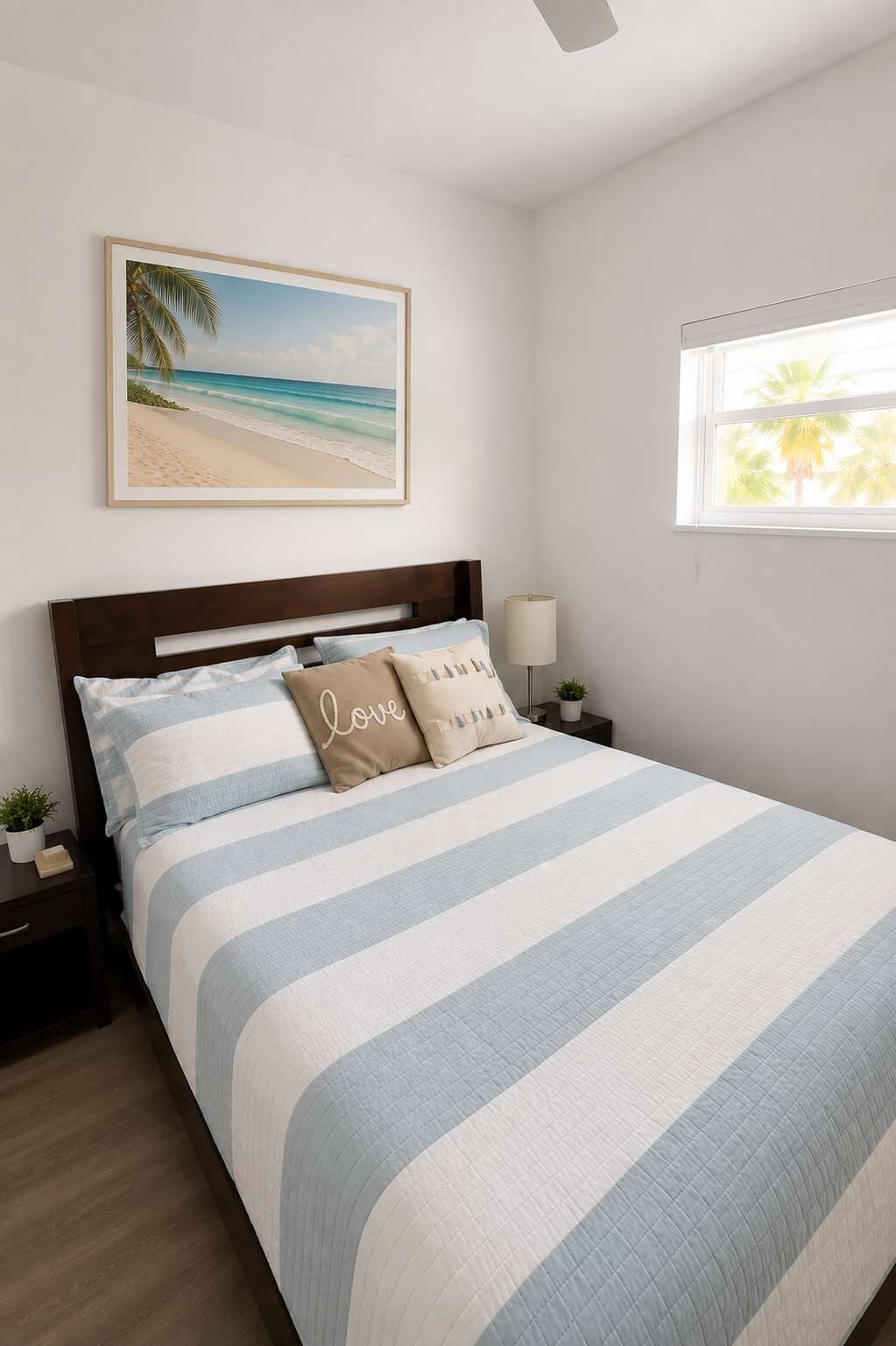 Ein Schlafzimmer mit einem Bett, einem Nachttisch, einer Lampe, einem gerahmten Bild von einem Strand und einem Fenster mit Blick auf Palmen.