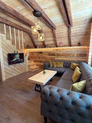Un salon chaleureux avec un canapé d’angle, un téléviseur à écran plat, des poutres en bois et un luminaire suspendu en forme d’étoile.