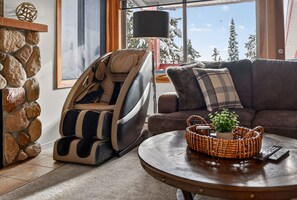 Un salon chaleureux avec un mur en pierre, un fauteuil massant, un canapé, une table basse agrémentée d’une plante, et une vue sur des arbres couverts de neige.