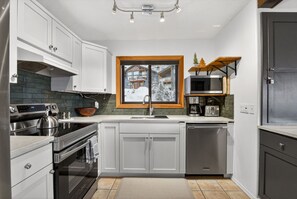 A modern kitchen with white cabinets, a stainless steel stove, a wooden bowl, and a window with a view of a house. - Big White Ski-In/Ski-Out Townhome | Private Hot Tub + Mountain Views (Kootenay Boundary E / West Bo*)