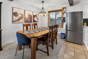 A dining area with a wooden table, chairs, and a rug. There is a kitchen with a refrigerator and cabinets. - Big White Ski-In/Ski-Out Townhome | Private Hot Tub + Mountain Views (Kootenay Boundary E / West Bo*)