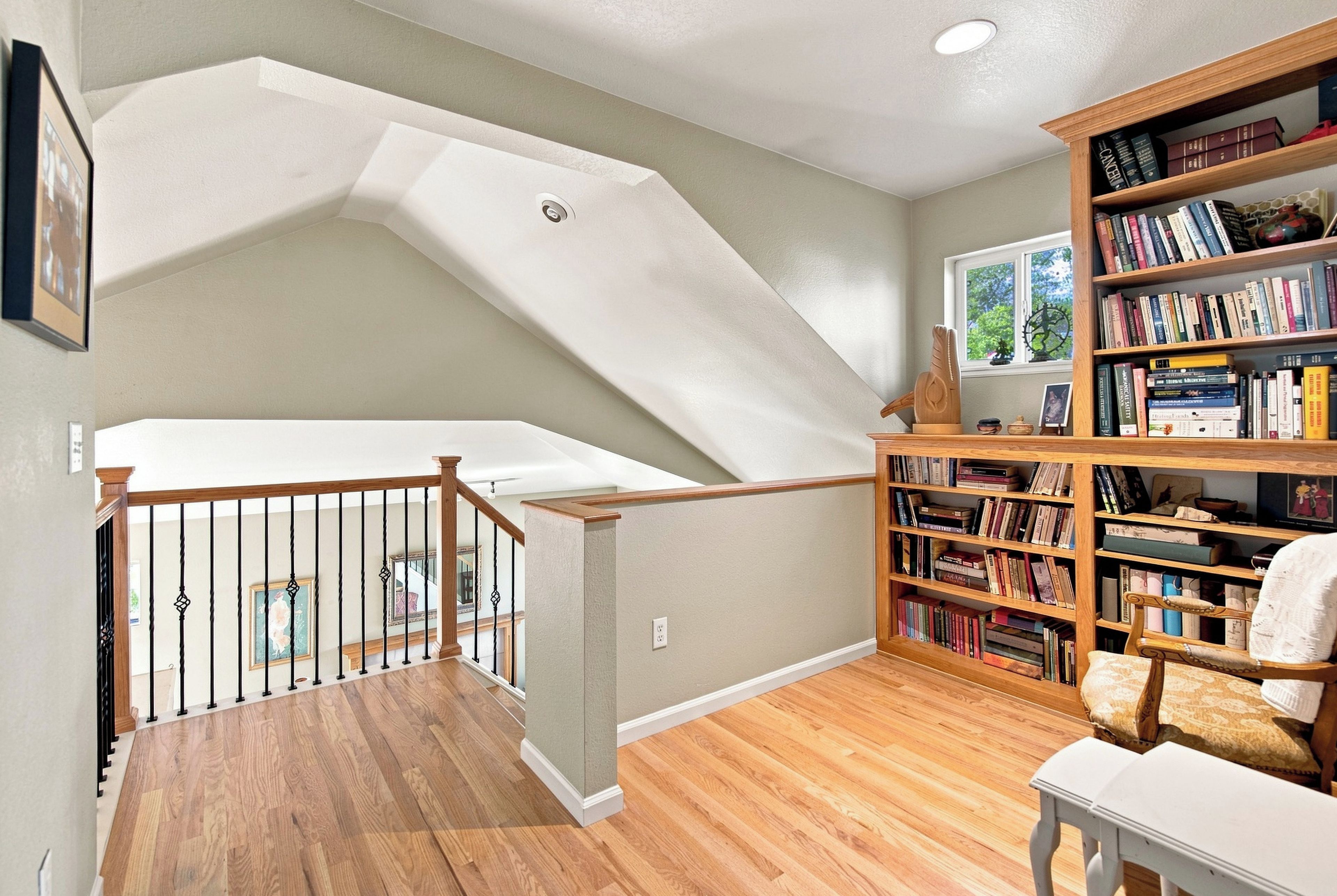 A spacious room with a wooden staircase, a bookshelf filled with books, and a window with a view of greenery.