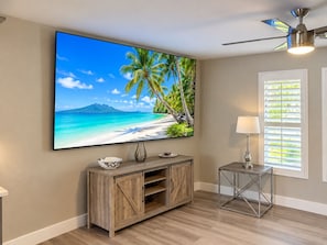 A room with a large flat-screen TV mounted on the wall, a wooden cabinet, a metal-framed side table, and a ceiling fan. -  Welcome to The Tropics (Cape Coral)
