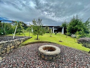 A stone fire pit surrounded by gravel, a swing set, and a building with a covered patio area.