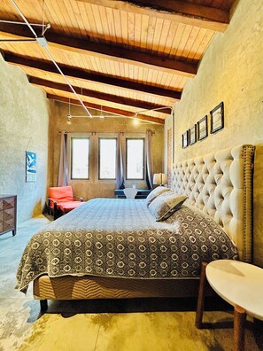 A bedroom with a large bed, a desk, and a chair. There are windows with curtains, a wooden ceiling, and a wall with framed pictures.