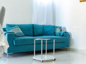 Ein hölzernes, rundes Beistelltisch steht neben einem smaragdgrünen Sofa mit gemusterten Kissen. Ein weißer Vorhang drapiert sich an einer weißen Wand.