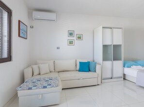 Ein moderner, minimalistischer Raum mit einem weißen Sofa, einem kleinen Hocker, einem Kleiderschrank und einem Bett mit einem blauen Kissen.