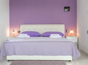 A bed with a white headboard, two purple pillows, and a white blanket. There are two bedside tables with lamps. - One bedroom apartment with Sea view and Terrace (5) (Podgora)