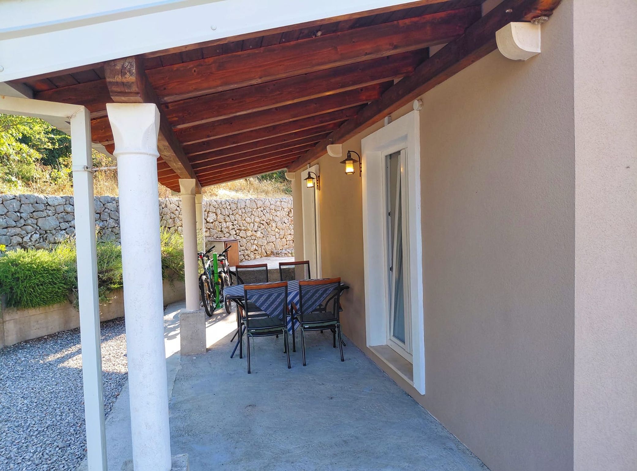 An outdoor patio area with a wooden pergola, a table with chairs, and a stone wall in the background.