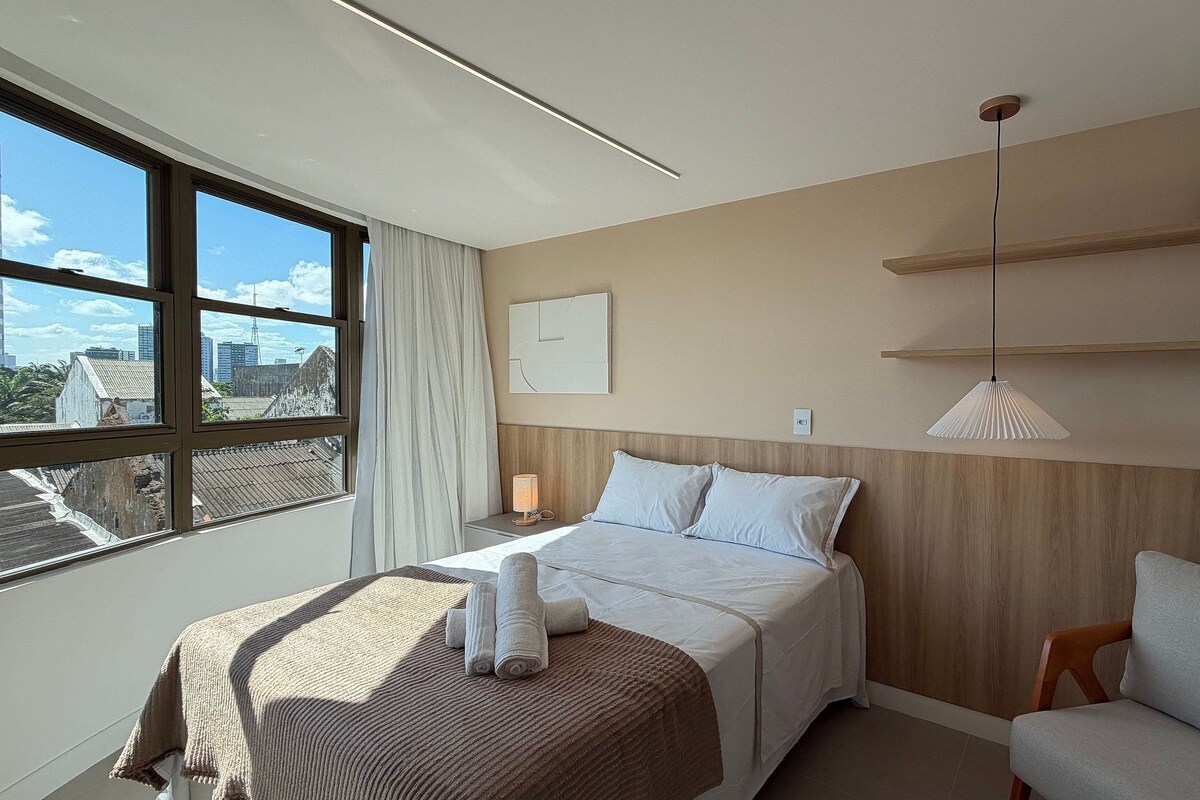 Habitación de hotel con cama, una silla, una ventana con vista a edificios y un estante en la pared con objetos decorativos.