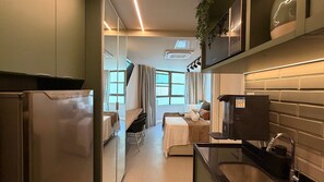 Habitación de hotel compacta con kitchenette, una cama con cabecera, una pequeña mesa de comedor y un ventanal que proporciona luz natural.