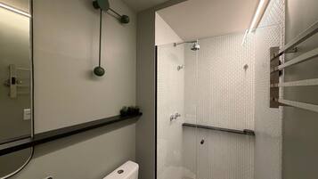 A bathroom with a glass shower enclosure, a toilet, a shelf with plants, and a mirror with a light fixture.
