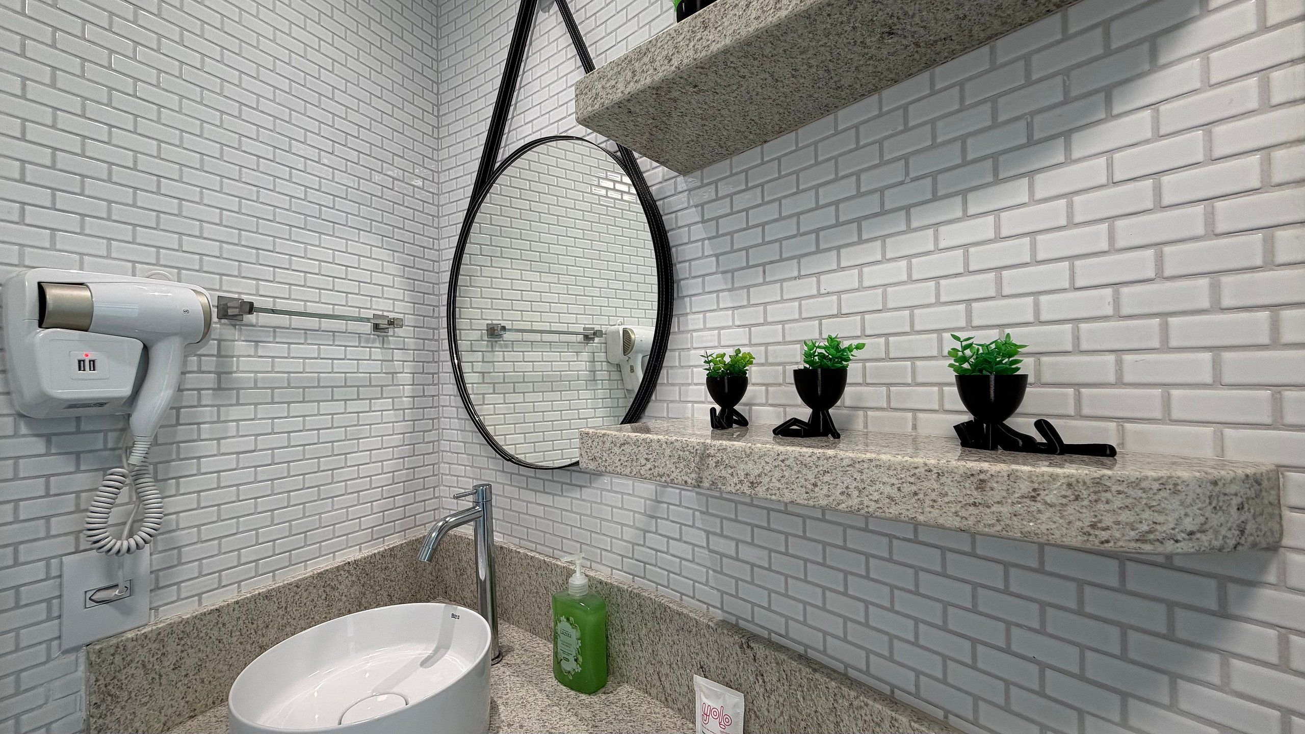 A bathroom with white brick walls, a white sink, a hairdryer, a mirror, and three black plant pots on a granite shelf.