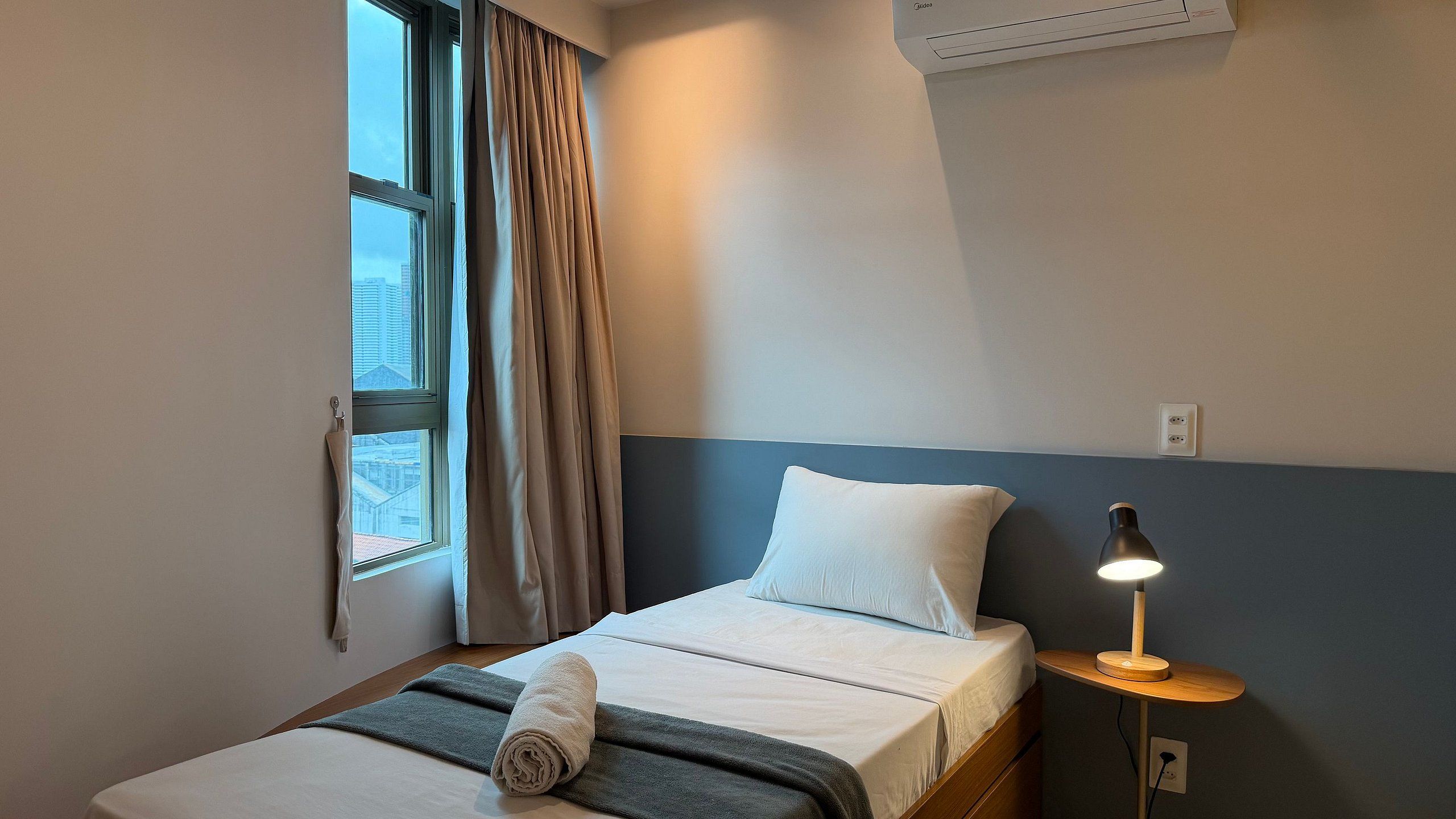 Una habitación de hotel con una cama, una mesita de noche con lámpara, una ventana con cortinas y un equipo de aire acondicionado.