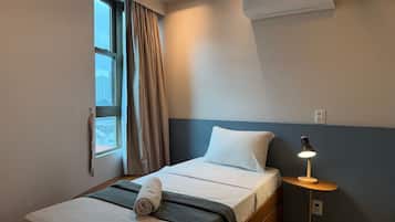 Una habitación de hotel con una cama, una mesita de noche con lámpara, una ventana con cortinas y un equipo de aire acondicionado.