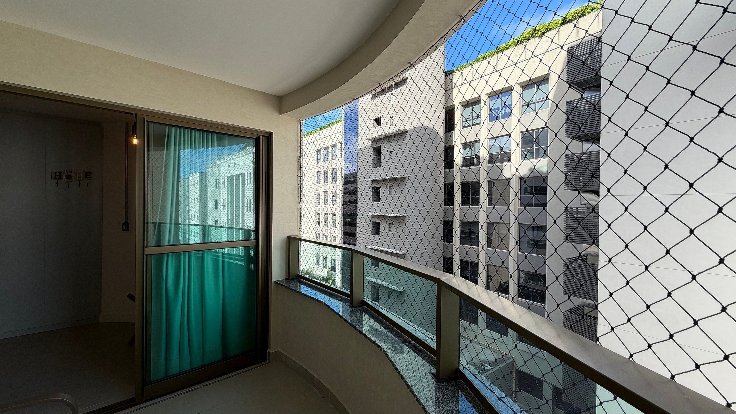 Un balcón con barandilla de alambre, una puerta de cristal y vistas a edificios cercanos.